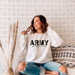 May include: White sweatshirt with the text "ARMY girlfriend" in black and camouflage print. The sweatshirt is long-sleeved and has a crew neck. The model is wearing blue jeans.