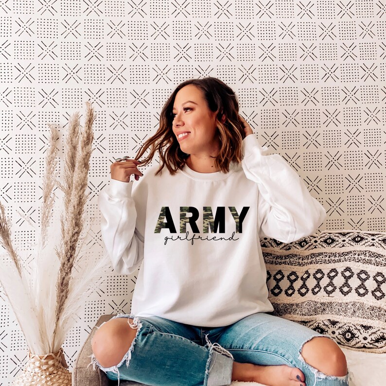 May include: White sweatshirt with the text "ARMY girlfriend" in black and camouflage print. The sweatshirt is long-sleeved and has a crew neck. The model is wearing blue jeans.