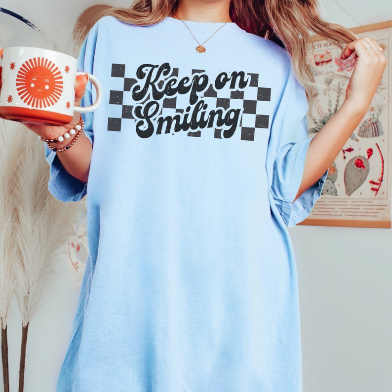 May include: A light blue oversized t-shirt with a black and white checkered pattern and the text "Keep on Smiling" in a retro font.