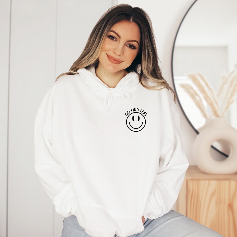 Puede incluir: Sudadera con capucha blanca con una carita sonriente negra y el texto "GO FIND LESS" en la parte delantera.