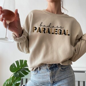 May include: A beige sweatshirt with the text "badass PARALEGAL" printed in black with a leopard print pattern.