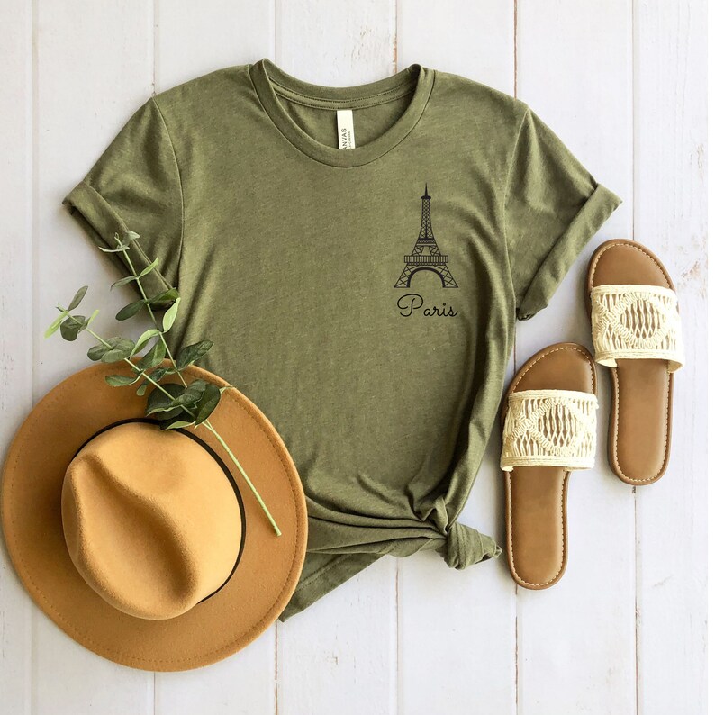 May include: A green t-shirt with a black outline of the Eiffel Tower and the word "Paris" underneath. The shirt is laid out on a white wooden surface with a brown hat and two pairs of sandals.