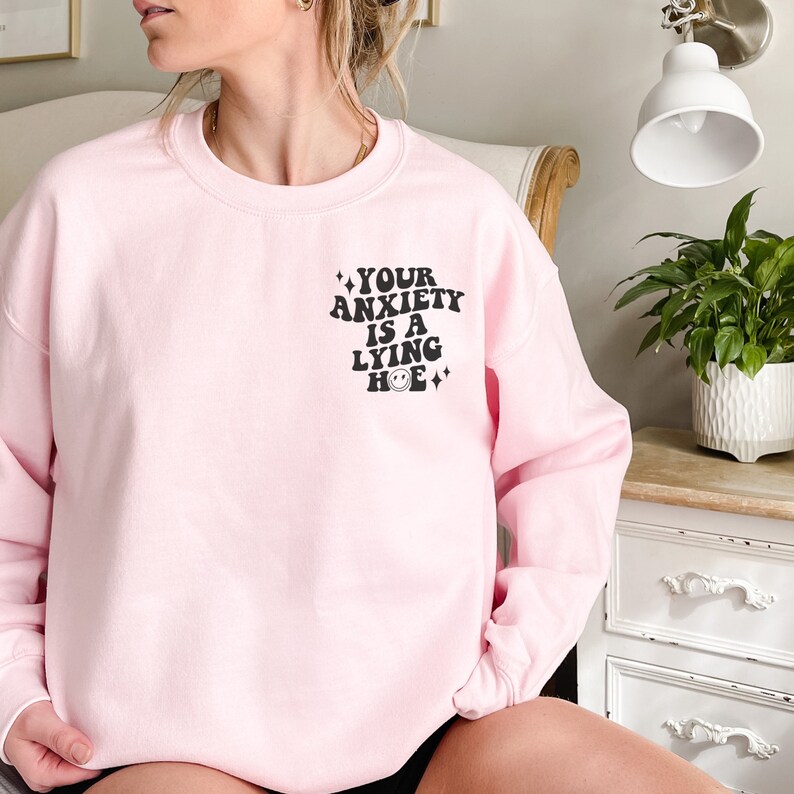 May include: Pink sweatshirt with the text "YOUR ANXIETY IS A LYING HOE" in black with a smiley face graphic.