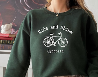 Puede incluir: Una sudadera verde oscuro con un gráfico blanco de una bicicleta y el texto "Ride and Shine Cycopath".