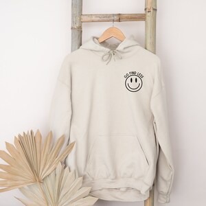 Puede incluir: Una sudadera con capucha beige con una carita sonriente negra y el texto "GO FIND LESS" en la parte delantera.