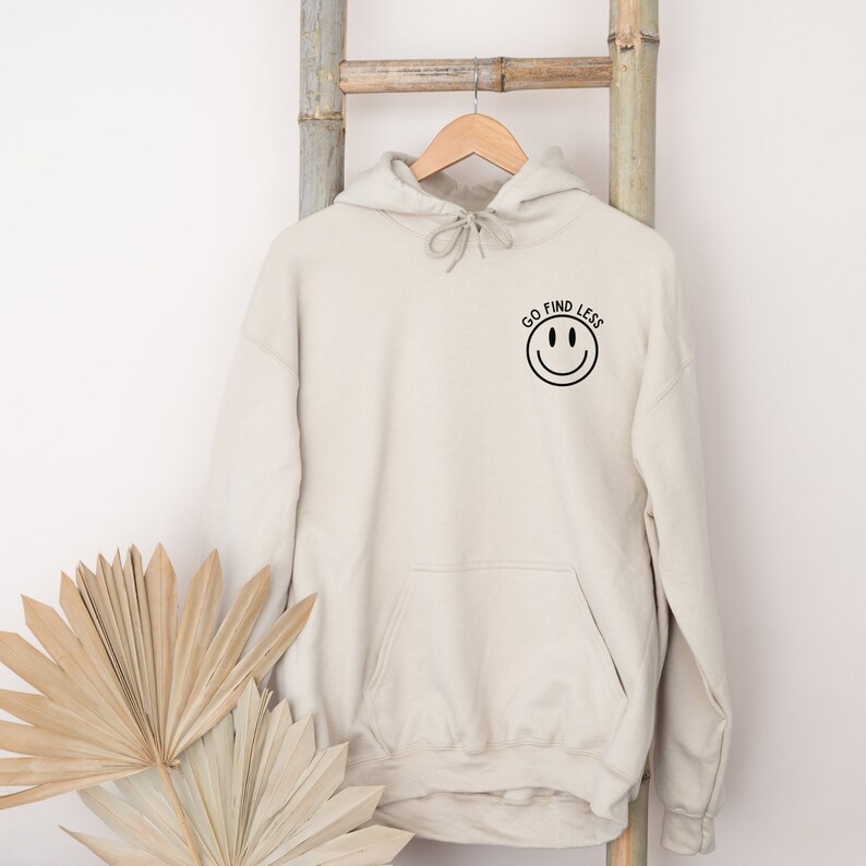 Puede incluir: Una sudadera con capucha beige con una carita sonriente negra y el texto "GO FIND LESS" en la parte delantera.