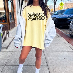 May include: A yellow oversized t-shirt with a black and white checkered pattern and the text "Keep on Smiling" in a retro font.