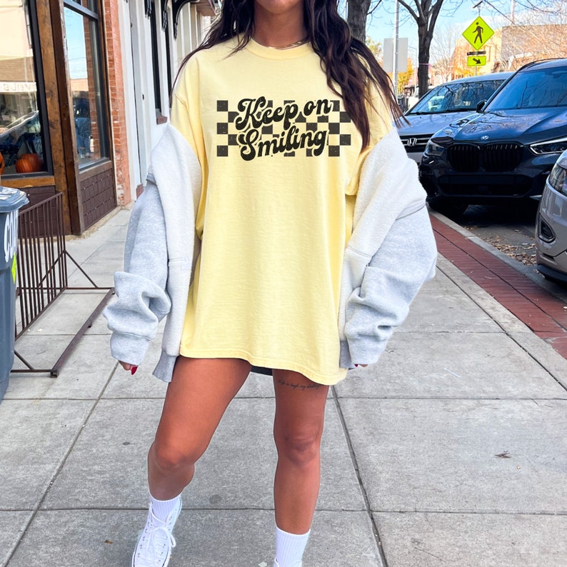 May include: A yellow oversized t-shirt with a black and white checkered pattern and the text "Keep on Smiling" in a retro font.