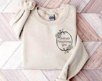 Puede incluir: Una sudadera beige con un estampado gráfico negro que dice "Obstinate Headstrong Girl" dentro de un círculo con detalles florales.