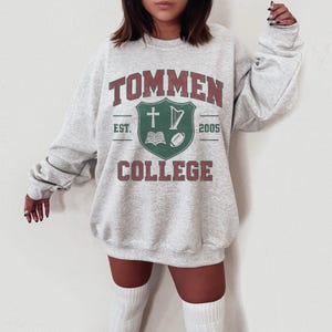Puede incluir: Una sudadera gris claro con el texto "TOMMEN COLLEGE" en rojo y verde. El diseño incluye un escudo con una cruz, arpa, libro y balón de fútbol. La sudadera tiene cuello redondo y mangas largas.