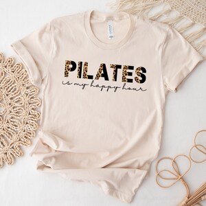 Op de afbeelding: Een beige T-shirt met de tekst "PILATES is my happy hour" in zwarte letters. Het woord "PILATES" is gedrukt in een luipaardprint.