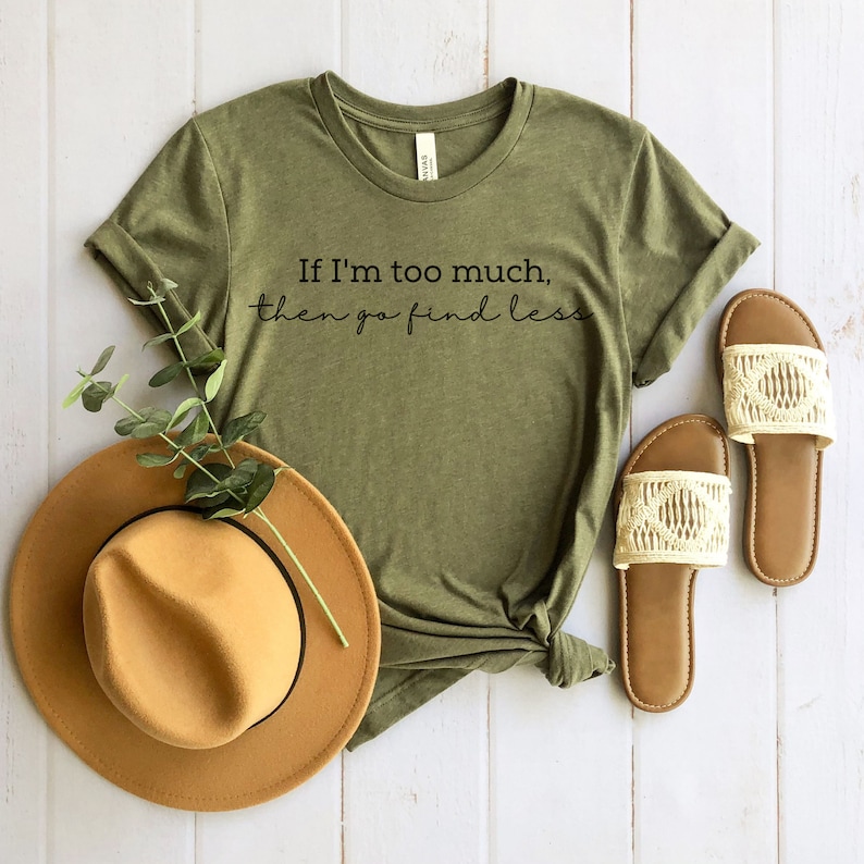 Op de afbeelding: Een groen T-shirt met de tekst "If I'm too much, then go find less" gedrukt op de voorkant. Het shirt ligt op een wit houten oppervlak met een bruine hoed en witte sandalen.