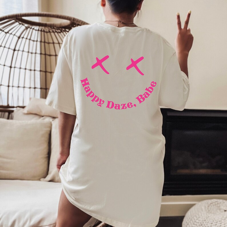 May include: White oversized t-shirt with a pink smiley face graphic and the text "Happy Daze, Babe" on the back.
