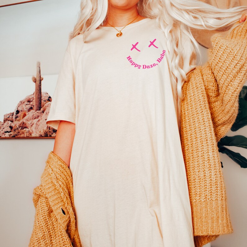 May include: A white t-shirt with a pink smiley face graphic and the text "Happy Daze, Babe" printed on the front. The shirt is being worn by a person with long blonde hair.