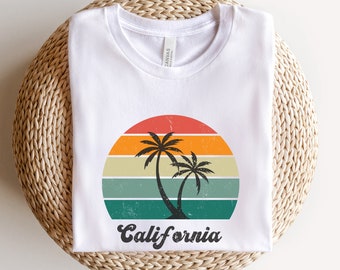 Op de afbeelding: Wit T-shirt met een vintage-stijl grafische afbeelding van palmbomen en een zonsondergang met de tekst "California" eronder.