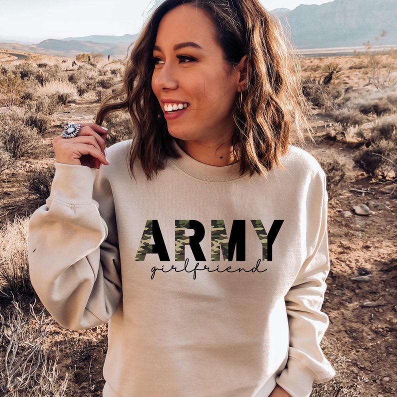 May include: A beige sweatshirt with the words "ARMY girlfriend" printed on the front. The word "ARMY" is in a combination of black and camouflage green letters. The word "girlfriend" is in a black script font.