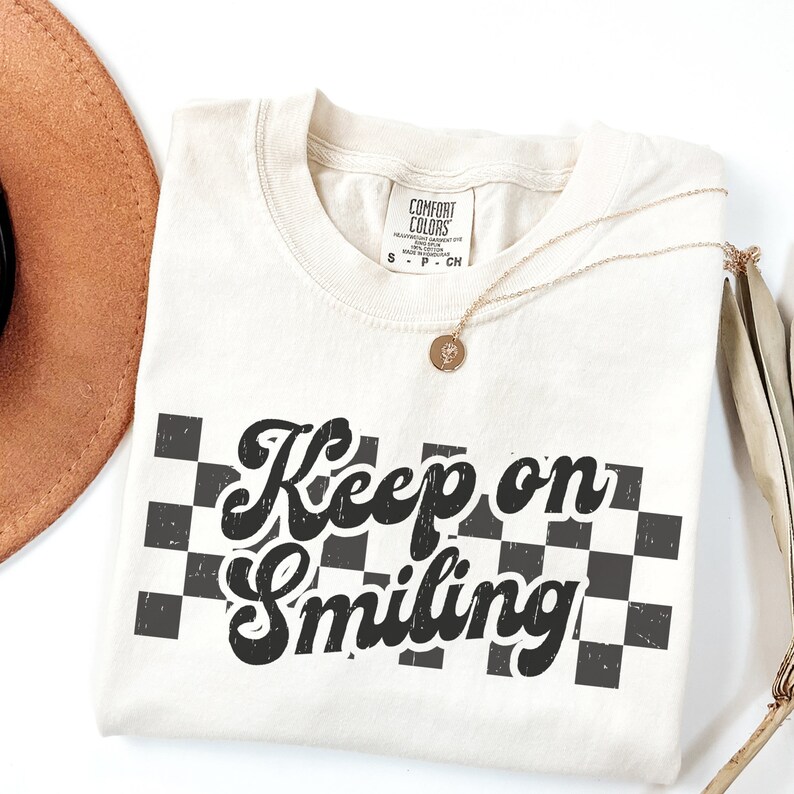 May include: A white t-shirt with a black and white checkered pattern and the text "Keep on Smiling" in a retro font.