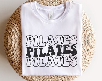 May include: White t-shirt with the text "PILATES" repeated three times in a retro font style. The text is black and the shirt is on a woven straw surface.