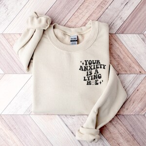 May include: A beige sweatshirt with the text "YOUR ANXIETY IS A LYING HOE" in black with a smiley face and sparkles.