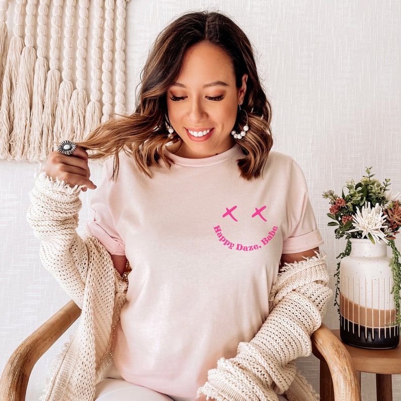 May include: A pink t-shirt with a pink smiley face and the text "Happy Daze, Babe" printed on the front.