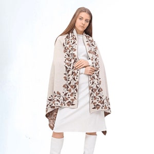 Op de afbeelding: Een beige sjaal met een bruin bloemenpatroon. De sjaal is over de schouders van een persoon gedrapeerd en wordt gedragen met een witte jurk en witte laarzen.