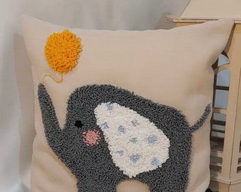 May include: A beige pillow with a gray elephant holding a yellow balloon. The elephant is made with a punch needle technique, creating a fluffy texture.
