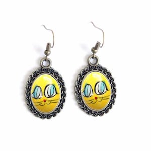 May include: A pair of dangle earrings with a yellow cat face design. The oval pendants have a black filigree border and are suspended from silver-coloured ear wires. The cat face has blue eyes, a red nose, and black whiskers.