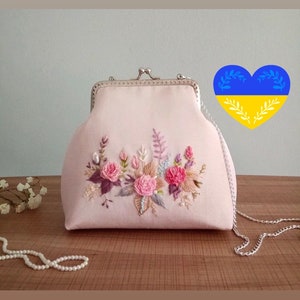 May include: A pink satin clutch bag with a floral embroidery design. The bag has a silver chain strap and a silver clasp.