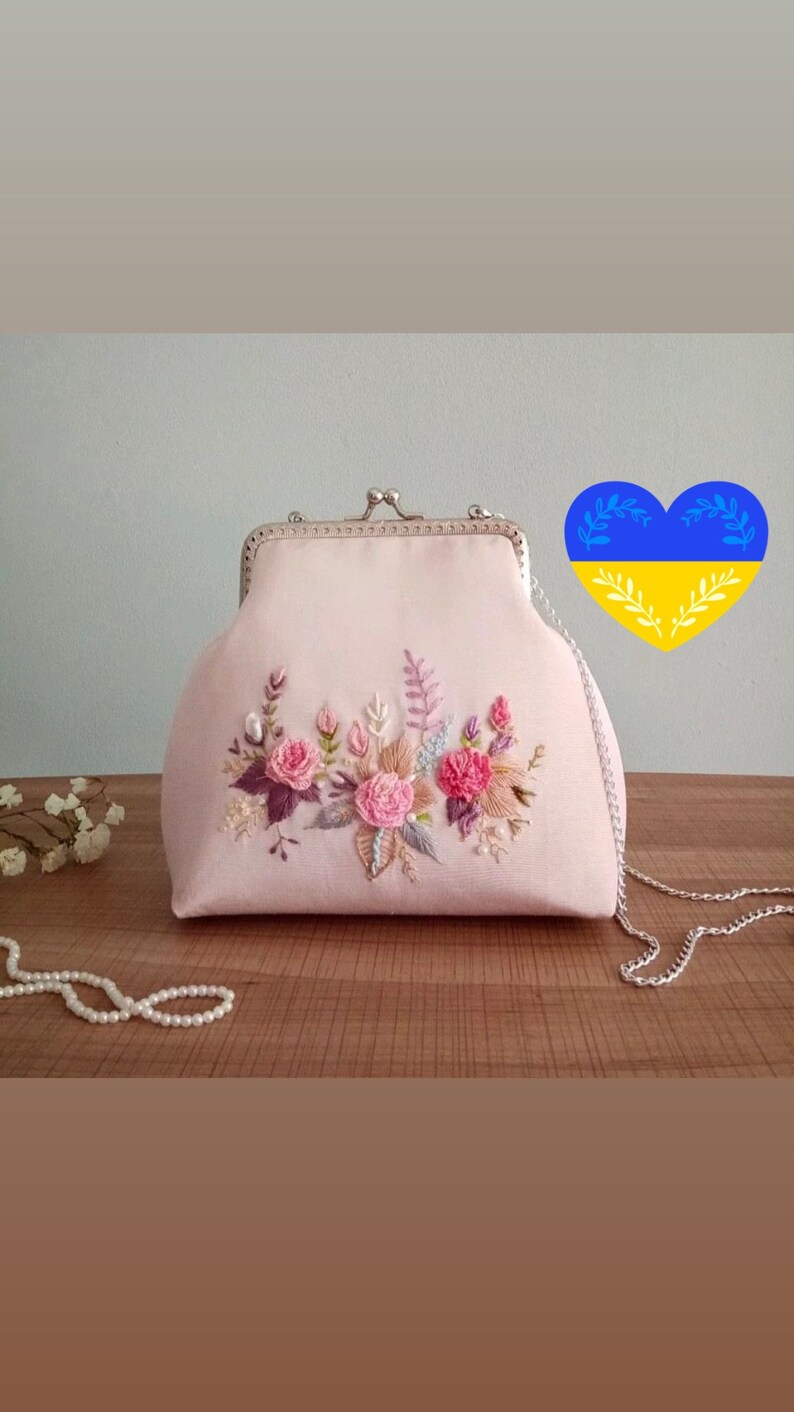 May include: A pink satin clutch bag with a floral embroidery design. The bag has a silver chain strap and a silver clasp.