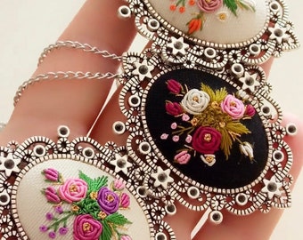 May include: Three silver oval-shaped pendants with intricate filigree designs. Each pendant features a different floral embroidery design on a white, black, and white background.