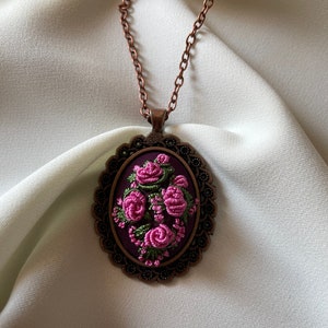 May include: A delicate necklace with a copper chain and an oval pendant featuring a floral embroidery design. The embroidery is done in shades of pink and green on a dark purple fabric. The pendant is framed by a decorative copper border.