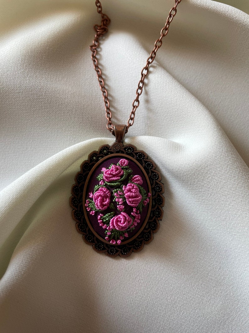 May include: A delicate necklace with a copper chain and an oval pendant featuring a floral embroidery design. The embroidery is done in shades of pink and green on a dark purple fabric. The pendant is framed by a decorative copper border.