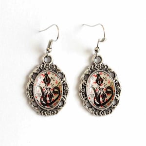 May include: A pair of silver earrings with a white oval pendant featuring a black and red floral design. The pendant is surrounded by a silver filigree frame.