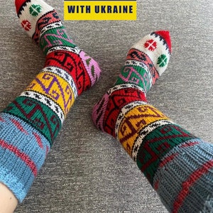 Könnte beinhalten: Ein Paar bunte Stricksocken mit einem blauen, roten, grünen, gelben und weißen Muster. Die Socken haben ein traditionelles ukrainisches Design. Der Text "SOLIDARITY WITH UKRAINE" wird im oberen Teil des Bildes angezeigt.