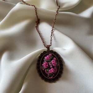 May include: A delicate necklace with a copper chain and an oval pendant featuring a floral embroidery design. The pendant is decorated with pink and green thread roses on a dark purple background.