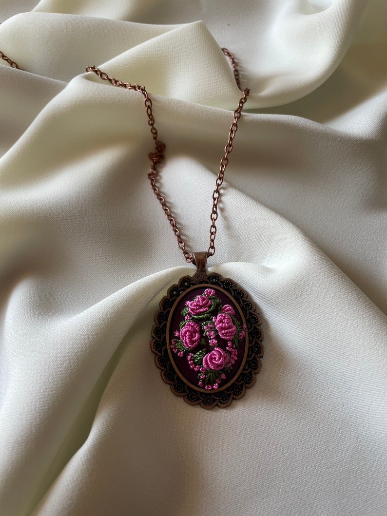 May include: A delicate necklace with a copper chain and an oval pendant featuring a floral embroidery design. The pendant is decorated with pink and green thread roses on a dark purple background.