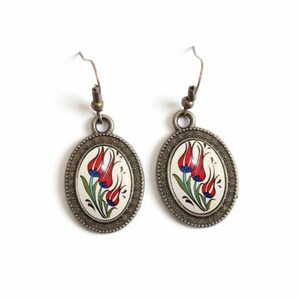 May include: A pair of oval-shaped earrings with a white background and a red, blue, and green floral design. The earrings have a bronze finish.