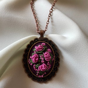 May include: A copper chain necklace with a purple and green embroidered floral design in an oval frame.