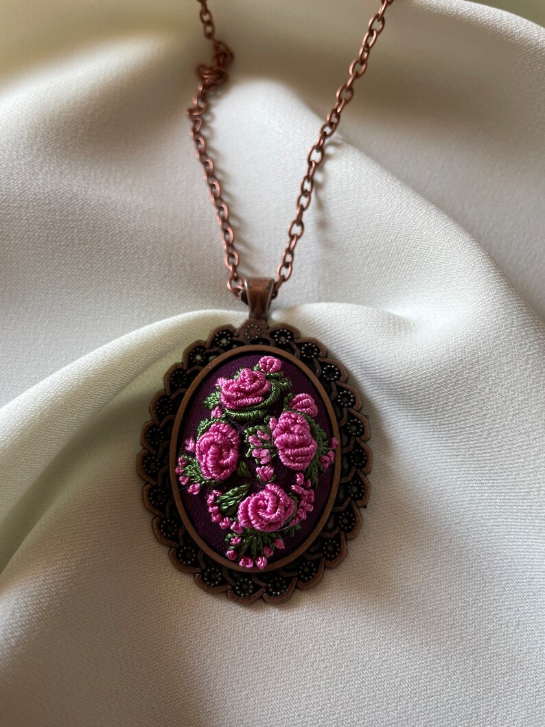 May include: A copper chain necklace with a purple and green embroidered floral design in an oval frame.