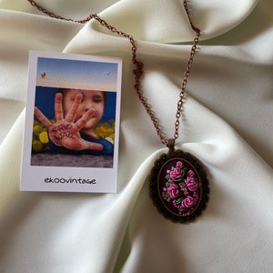 May include: A copper chain necklace with an oval pendant featuring a floral embroidery design. The pendant is surrounded by a decorative metal frame. The embroidery is in shades of pink and green on a dark purple background.