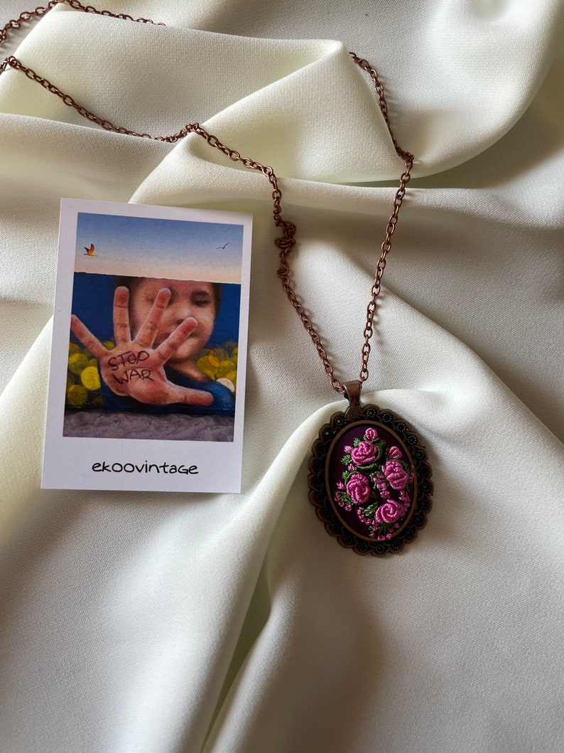 May include: A copper chain necklace with an oval pendant featuring a floral embroidery design. The pendant is surrounded by a decorative metal frame. The embroidery is in shades of pink and green on a dark purple background.
