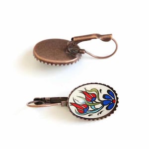 May include: A pair of copper-toned earrings with a white oval-shaped ceramic charm featuring a blue and red floral design.
