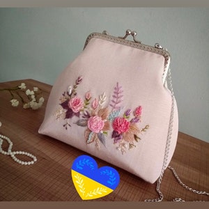 May include: A pink fabric clutch bag with a silver clasp and chain strap. The bag is decorated with embroidered flowers in shades of pink, purple, and brown.