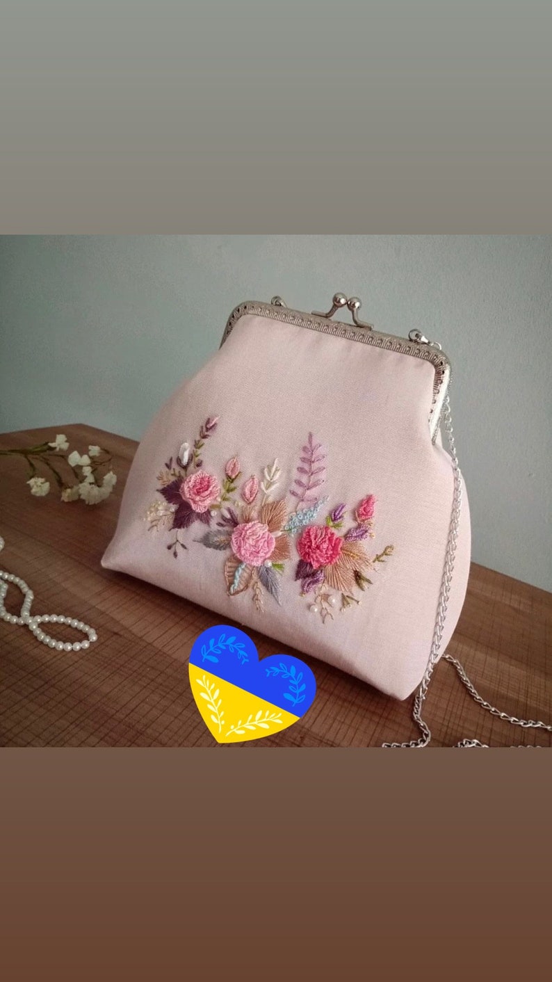 May include: A pink fabric clutch bag with a silver clasp and chain strap. The bag is decorated with embroidered flowers in shades of pink, purple, and brown.