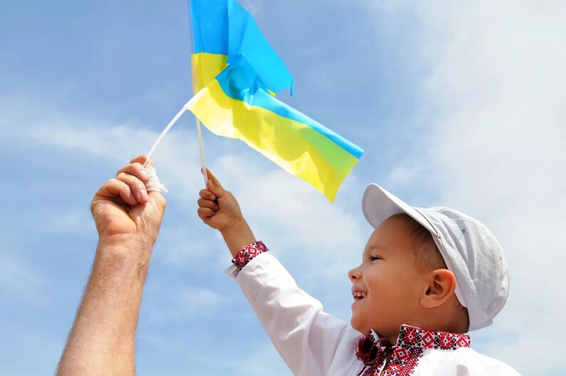 May include: A young boy wearing a white shirt and a white cap holds a blue and yellow flag with a man's hand holding the other end of the flag.