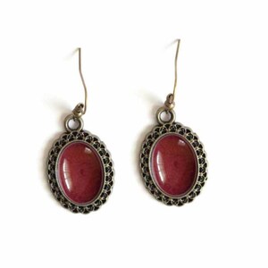 May include: A pair of oval-shaped earrings with a burgundy red enamel inlay. The earrings have a delicate, ornate, gold-toned metal frame.