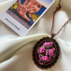 May include: A copper chain necklace with a pendant featuring a purple oval cameo with pink embroidered roses. The cameo is surrounded by a decorative metal frame.
