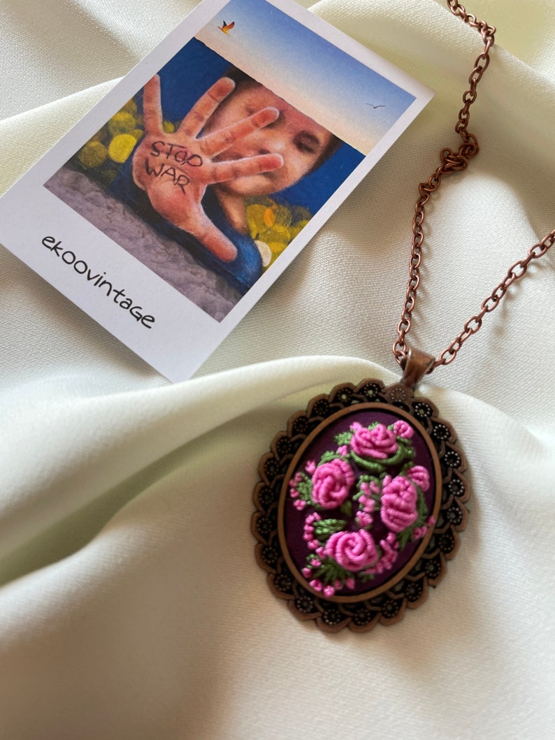 May include: A copper chain necklace with a pendant featuring a purple oval cameo with pink embroidered roses. The cameo is surrounded by a decorative metal frame.