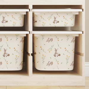 Pode incluir: Uma unidade de armazenamento de madeira clara com seis cubículos, cada um com um recipiente creme. Os recipientes apresentam um padrão repetido de coelhos, flores e folhas. A unidade está sobre um chão de madeira clara.