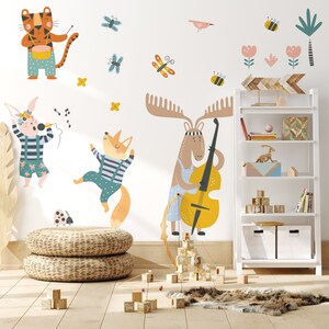 Puede incluir: Un conjunto de pegatinas de pared coloridas con un tigre tocando la batería, un zorro tocando la guitarra, un alce tocando el violonchelo y un conejo cantando. Las pegatinas están en una pared blanca con un estante blanco y bloques de madera en primer plano.